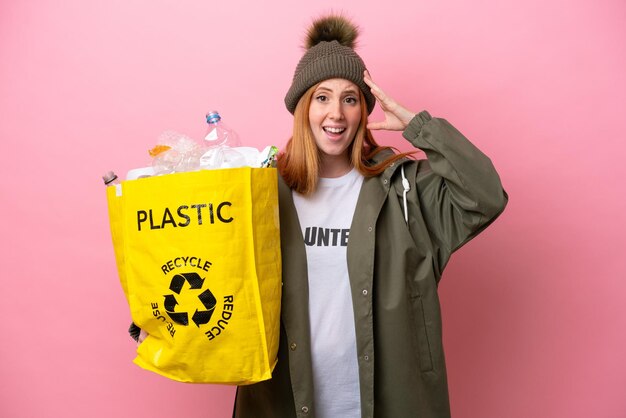 Mulher jovem ruiva segurando um saco cheio de garrafas plásticas para reciclar isolado em fundo rosa com expressão de surpresa