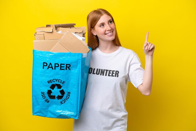 Mulher jovem ruiva segurando um saco de reciclagem cheio de papel para reciclar isolado em fundo amarelo apontando uma ótima ideia