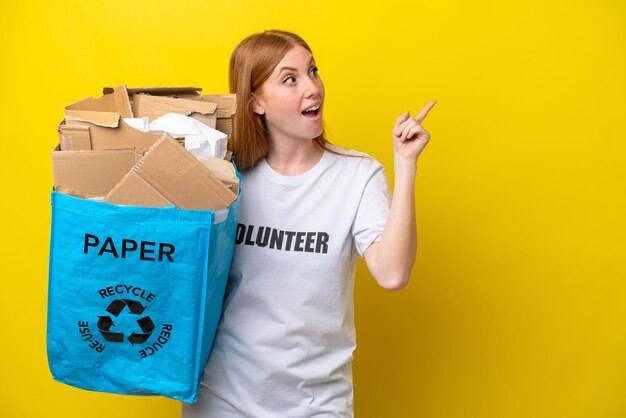 Mulher jovem ruiva segurando um saco de reciclagem cheio de papel para reciclar isolado em fundo amarelo com a intenção de perceber a solução enquanto levanta um dedo