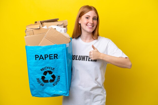 Mulher jovem ruiva segurando um saco de reciclagem cheio de papel para reciclar isolado em fundo amarelo, dando um polegar para cima gesto