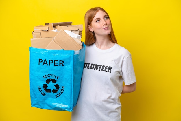 Mulher jovem ruiva segurando um saco de reciclagem cheio de papel para reciclar isolado em fundo amarelo e olhando para cima