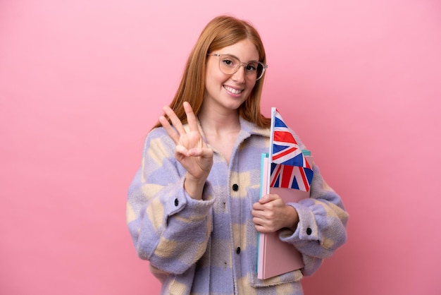 Mulher jovem ruiva segurando uma bandeira do Reino Unido isolada em fundo rosa feliz e contando três com os dedos