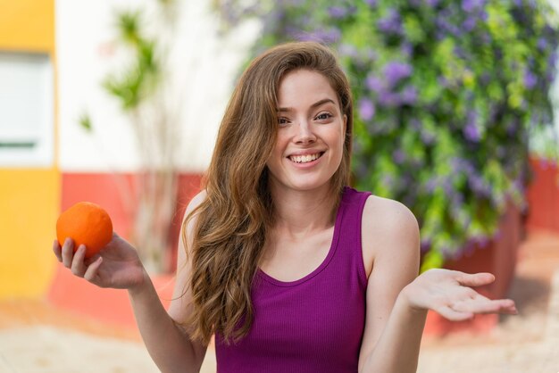 Mulher jovem ruiva segurando uma laranja ao ar livre com expressão facial chocada