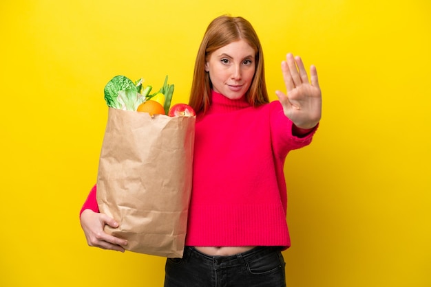 Mulher jovem ruiva segurando uma sacola de compras isolada em fundo amarelo, fazendo o gesto de parada