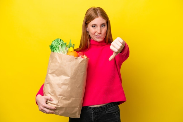 Mulher jovem ruiva segurando uma sacola de compras isolada em fundo amarelo, mostrando o polegar para baixo com expressão negativa