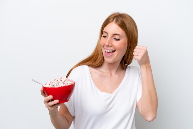 Mulher jovem ruiva segurando uma tigela de cereais isolada no fundo branco comemorando uma vitória