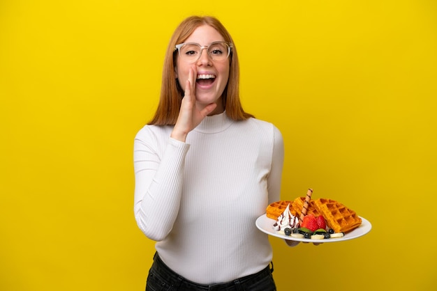 Mulher jovem ruiva segurando waffles isolados em fundo amarelo gritando com a boca aberta