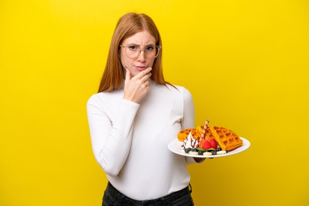 Mulher jovem ruiva segurando waffles isolados no pensamento de fundo amarelo