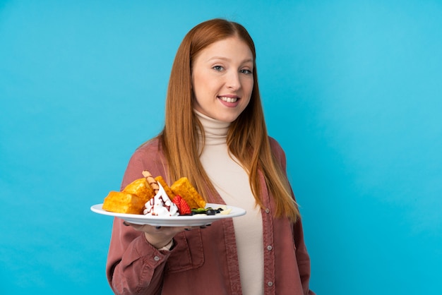 Mulher jovem ruiva segurando waffles sobre parede isolada com expressão feliz