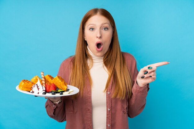 Mulher jovem ruiva segurando waffles sobre parede isolada surpreendeu e apontando o lado