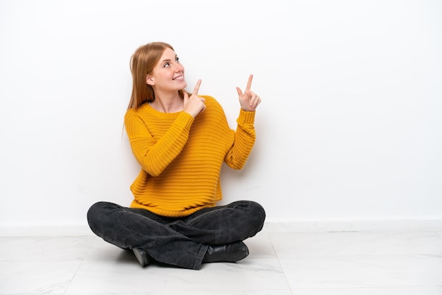 Mulher jovem ruiva sentada no chão isolada no fundo branco apontando com o dedo indicador uma ótima ideia