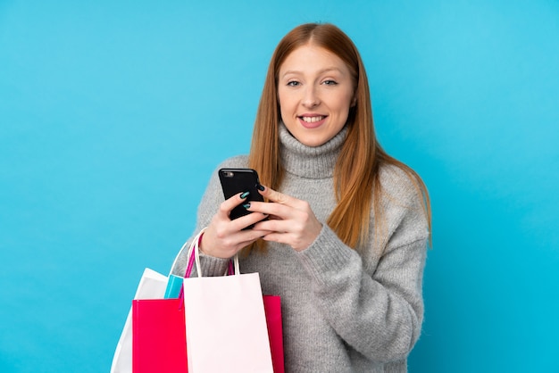 Mulher jovem ruiva sobre parede azul isolada segurando sacolas de compras e escrever uma mensagem com o celular para um amigo