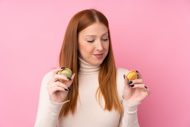 Mulher jovem ruiva sobre parede rosa isolada segurando macarons franceses coloridos e olhando