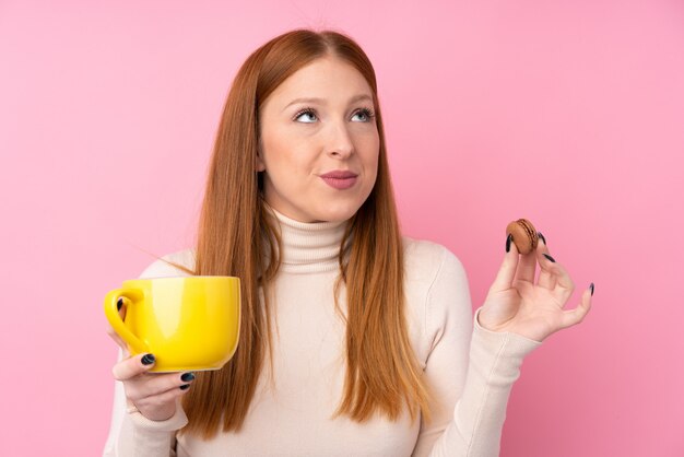 Mulher jovem ruiva sobre parede rosa isolada segurando macarons franceses coloridos e um copo de leite