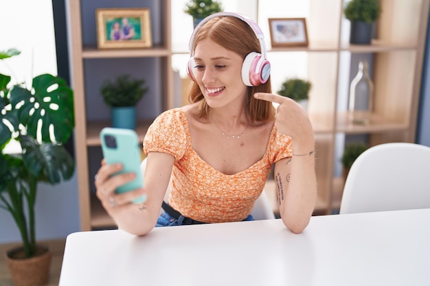 Mulher jovem ruiva usando smartphone fazendo videochamada sentado na mesa sorrindo feliz apontando com a mão e o dedo