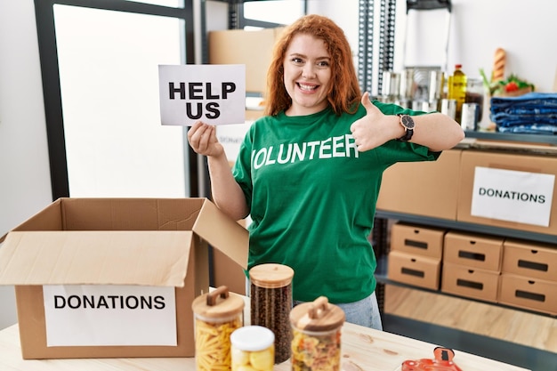 Mulher jovem ruiva vestindo camiseta de voluntário segurando ajuda nos banner sorrindo feliz e positivo polegar para cima fazendo excelente e sinal de aprovação