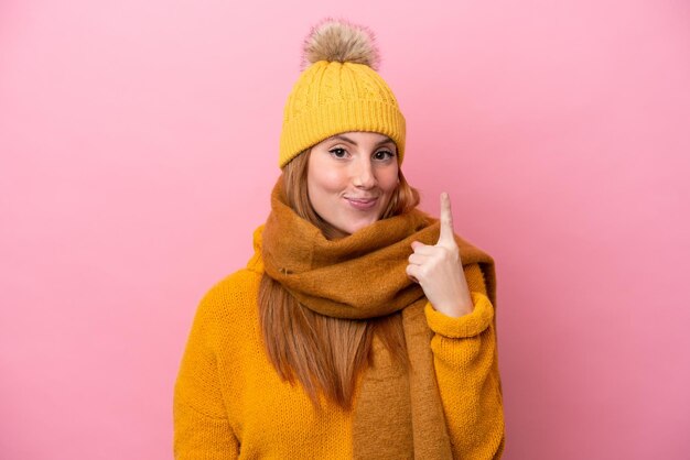 Mulher jovem ruiva vestindo jaqueta de inverno isolada em fundo rosa apontando com o dedo indicador uma ótima idéia