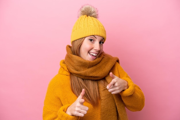 Mulher jovem ruiva vestindo jaqueta de inverno isolada em fundo rosa, apontando para a frente e sorrindo