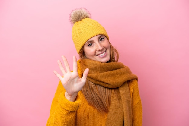 Mulher jovem ruiva vestindo jaqueta de inverno isolada em fundo rosa contando cinco com os dedos