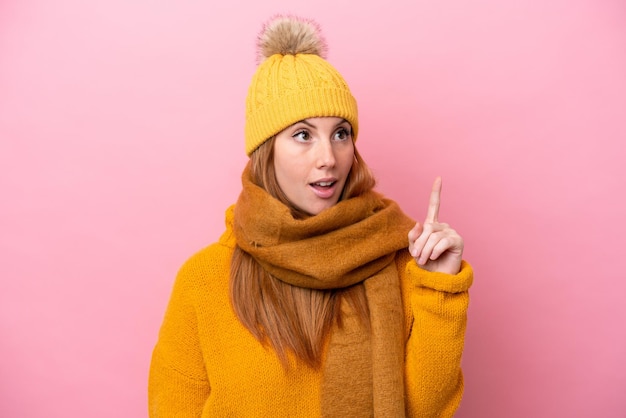 Mulher jovem ruiva vestindo jaqueta de inverno isolada em fundo rosa pensando em uma ideia apontando o dedo para cima