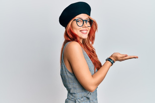 Mulher jovem ruiva vestindo moda francesa com boina apontando para o lado com as palmas das mãos abertas mostrando espaço para cópia, apresentando anúncio sorrindo animado feliz