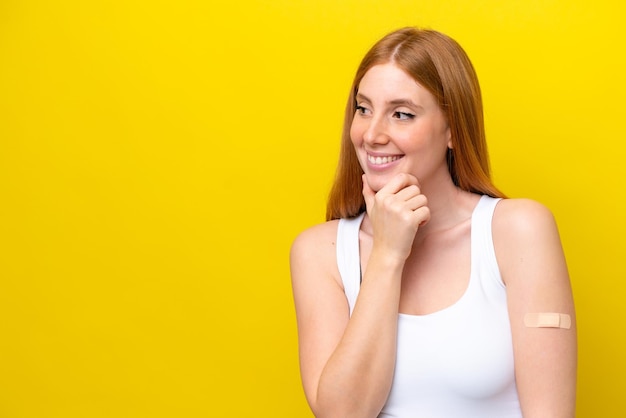 Mulher jovem ruiva vestindo um curativo isolado em fundo amarelo, pensando em uma ideia e olhando para o lado