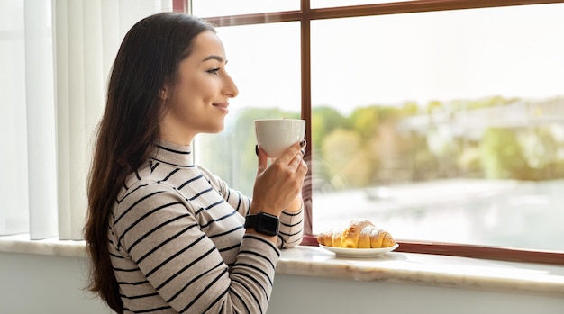 Foto mulher jovem satisfeita desfrutando de um momento de paz bebendo café e olhando pela janela ensolarada