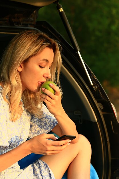 Mulher jovem se senta no porta-malas aberto de um carro e come uma maçã verde.