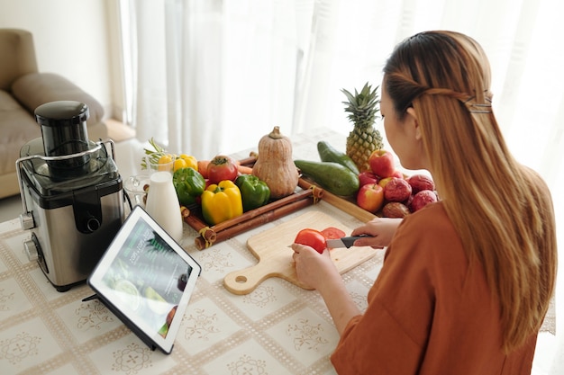 Mulher jovem seguindo a receita no computador tablet, ela está fatiando tomates com uma faca afiada na mesa da cozinha