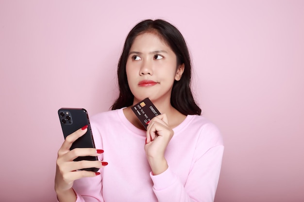 Mulher jovem segura um smartphone e um cartão de crédito em sua mão Mulher asiática posa com equipamentos em fundo rosa claro