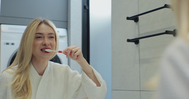 Foto mulher jovem segurando escova de dentes escovando dentes olhando para o espelho limpando a boca fazendo a rotina matinal de cuidados dentários orais no banheiro