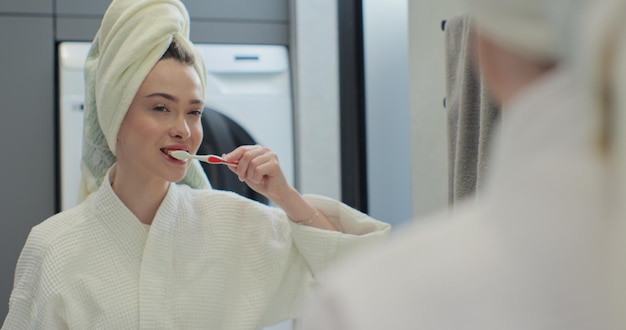 Mulher jovem segurando escova de dentes escovando dentes olhando para o espelho limpando a boca