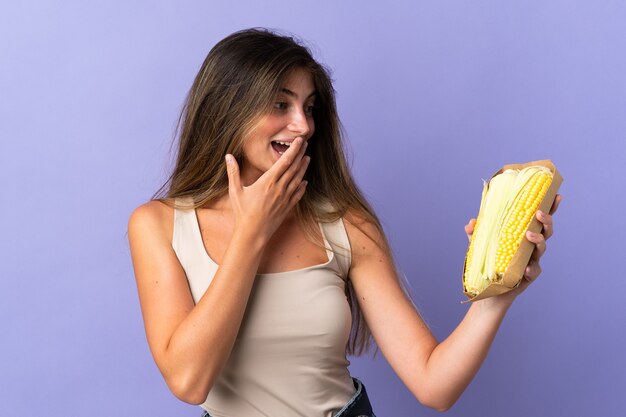 Mulher jovem segurando milho isolado no roxo com expressão facial surpresa e chocada