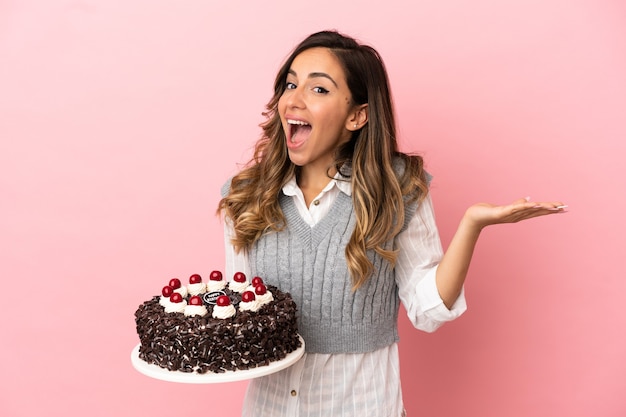 Mulher jovem segurando um bolo de aniversário sobre um fundo rosa isolado com uma expressão facial chocada