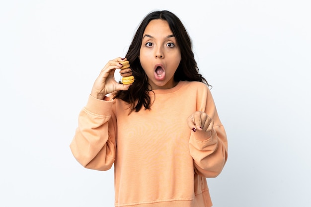 Mulher jovem segurando um fundo branco isolado segurando macarons franceses coloridos e se surpreendendo ao apontar para a frente