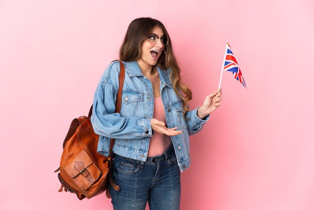 Mulher jovem segurando uma bandeira do Reino Unido isolada em rosa com expressão de surpresa enquanto olha para o lado