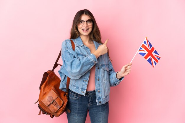 Mulher jovem segurando uma bandeira do Reino Unido isolada em uma parede rosa apontando para o lado para apresentar um produto