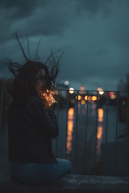 Foto mulher jovem segurando uma luz de corda enquanto estava sentada em uma parede de retenção sobre o rio contra o céu ao anoitecer