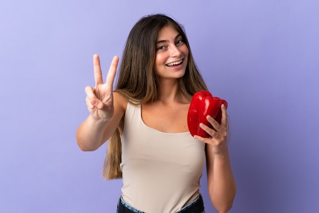 Mulher jovem segurando uma pimenta isolada na parede roxa sorrindo e mostrando o sinal da vitória