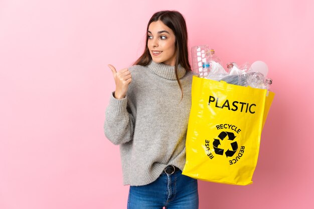 Mulher jovem segurando uma sacola cheia de garrafas de plástico para reciclar isolada em uma parede rosa apontando para o lado para apresentar um produto