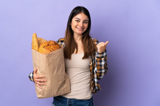 Mulher jovem segurando uma sacola cheia de pães isolada na parede roxa apontando para o lado para apresentar um produto