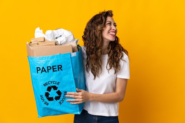 Mulher jovem segurando uma sacola cheia de papel para reciclar isolada na parede amarela rindo em posição lateral
