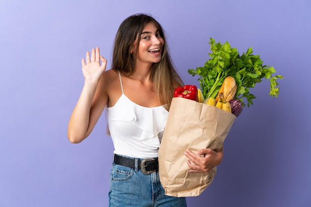 Mulher jovem segurando uma sacola de compras de supermercado e saudando com a mão com expressão feliz