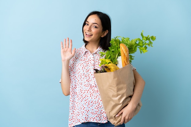 Mulher jovem segurando uma sacola de compras de supermercado e saudando com a mão com expressão feliz