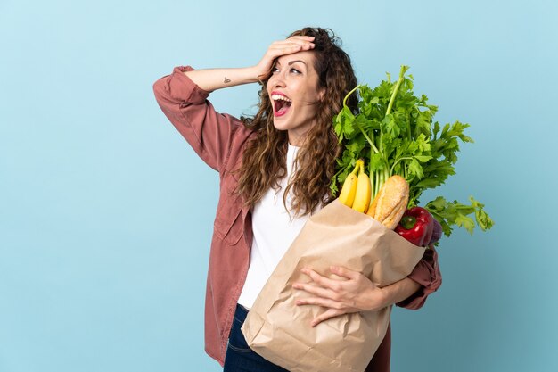Mulher jovem segurando uma sacola de compras de supermercado isolada em azul fazendo gesto de surpresa enquanto olha para o lado