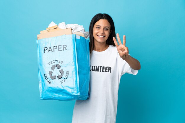 Mulher jovem segurando uma sacola de reciclagem cheia de papel feliz e contando três com os dedos