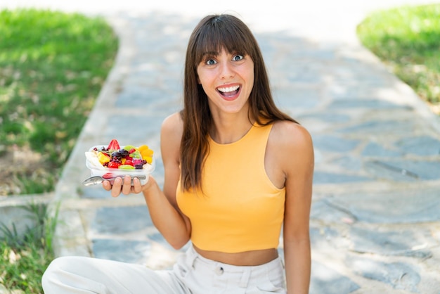 Mulher jovem segurando uma tigela de frutas ao ar livre com expressão facial surpresa e chocada