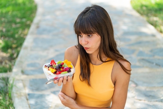 Mulher jovem segurando uma tigela de frutas ao ar livre com expressão triste