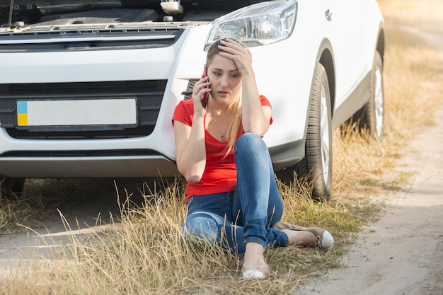 Mulher jovem sentada ao lado de um carro quebrado e pedindo ajuda