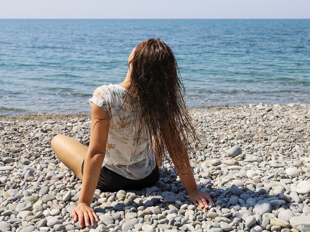 Mulher jovem sentada de costas na praia de seixos e sacudindo o cabelo molhado